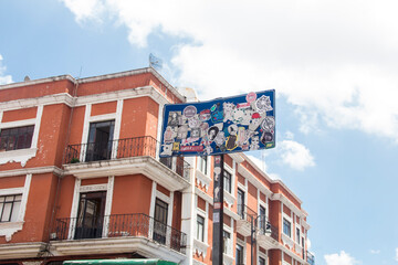 Public sign covered in colorful stickers with a historic Mexican colonial building in the background, creating a striking contrast between urban street art and classic architecture.