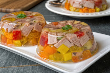 two dishes of a jellied meat and vegetable salad on a white plate, garnished with fresh herbs. The background includes a cutting board and a fork, suggesting a dining setting.