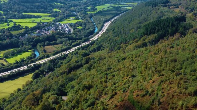 Aerial View Over Forest Covered Hill with Cluster of Residential Houses with Busy Dual Carriageway on A465 Heads of the Valleys Road. Winding River Neath in Background and Open Fields.