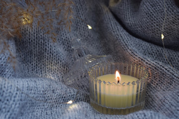 A lit candle on warm blanket decorated with garland. Cozy atmosphere with a candle. 