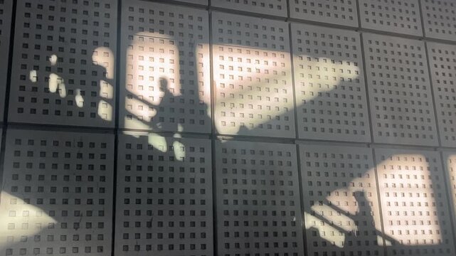 A fixed shot captures shadows of an escalator and people projected by sunlight on a patterned wall in a train station, creating a poetic and graphic scene.