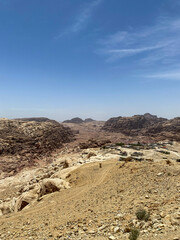 Wide-angle view of a desolate, rocky desert landscape in Jordan. The foreground is composed of dry, gravelly earth leading to rugged, light-colored rock formations under a clear blue sky.
