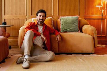 Smiling bearded man sitting on floor in cozy home interior, fashion model posing