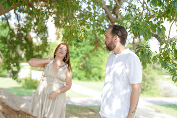 A man and a woman are standing under a tree. The man is wearing a white shirt and the woman is wearing a dress. Scene is casual and relaxed