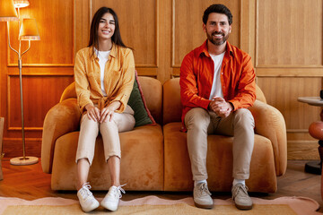 Obraz premium Smiling man and woman social distancing on sofa. Couple sitting in living room looking at camera