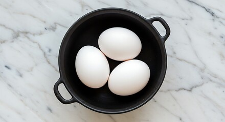 Three white eggs in a black bowl on a marble surface overhead view