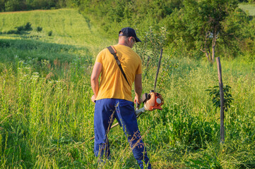 Man mowing tall grass with a gas-powered lawn mower in a rural field, surrounded by greenery, trees on a bright, sunny spring day
