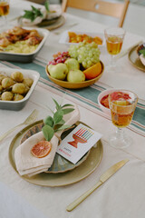 Closeup showing festive Kwanzaa table setting with card featuring kinara illustration, plate decorated with dried citrus slice and greenery, assorted fruit and food dishes arranged on table