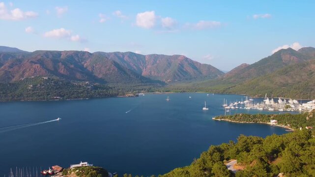 aerial perspective captures the serene beauty of Marmaris Bay in Turkey, featuring lush green mountains, a tranquil blue sea dotted with sailboats, and a bustling marina filled with yachts under a cle