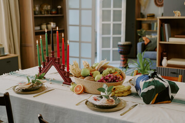 Kwanzaa table setting featuring kinara with seven candles, basket filled with assorted fruits and vegetables, traditional gifts and place settings arranged for celebration