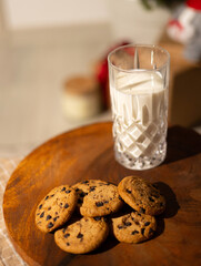Milk and cookies on wooden board near Christmas tree in soft light. Boy in pajamas prepares sweet treats and drink for Santa before bedtime. Cozy winter holiday atmosphere.