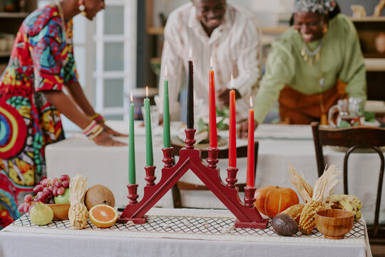 Three Black adults celebrating Kwanzaa, arranging festive table with kinara holding seven lit candles, assorted fruits, vegetables, and traditional decorations in foreground