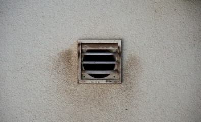 Close-up of a weathered metal ventilation grille on a textured exterior wall, showing signs of dust and age. Industrial detail highlighting airflow, building infrastructure, and urban decay.