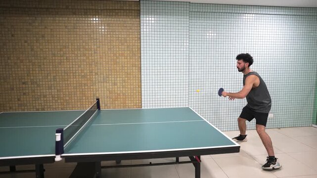 A left-handed player serves in an indoor table tennis match, captured in a static wide-angle shot. The frame highlights his athletic stance, controlled movement, and energetic lifestyle.