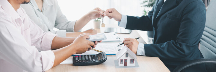 Panorama of real estate deal with house models, key handover, cash money, and calculator on desk, symbolizing property investment, mortgage loan, housing purchase, and financial planning success.