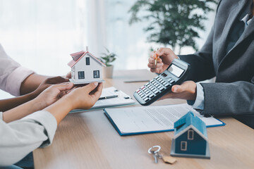 Close up of real estate agent handing house model to client with calculator and contract paperwork on desk, concept of mortgage loan, home ownership, property investment and housing agreement.