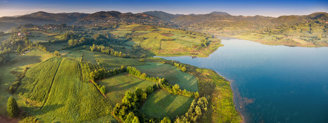 Rural countryside panorama with lush green fields, rolling hills, and a calm blue lake under a clear sky