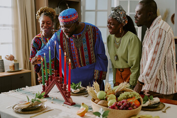 Group of Black adults and young adult woman celebrating Kwanzaa, lighting kinara candles together at table with traditional fruits and decorations, smiling and interacting