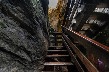Narrow alpine gorge with wooden walkway, dramatic rock formations and natural light, dark mountain canyon passage creating adventurous hiking atmosphere