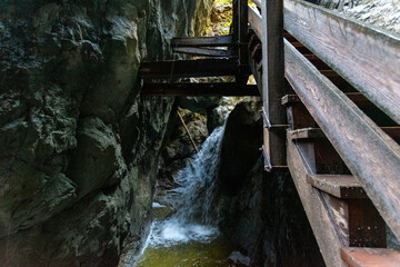 Narrow alpine gorge with wooden walkway, dramatic rock formations and natural light, dark mountain canyon passage creating adventurous hiking atmosphere