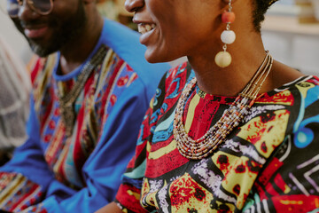 Fototapeta premium Black woman and Black man sitting together smiling and talking during Kwanzaa celebration, both wearing colorful patterned clothing and beaded jewelry, upper bodies visible