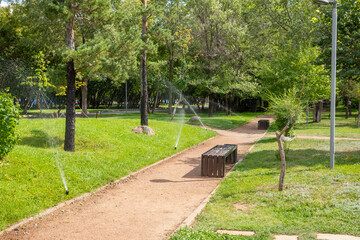 Sprinklers water lush green grass in peaceful park on sunny day. Walkways lined with benches. improvement, beautification, renovation of the urban environment, improving quality of life