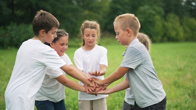 Children playing hand game in grass field with five kids forming circle placing palm together showing team and friend bond during outdoor play activity summer game promoting group bonding by tree line