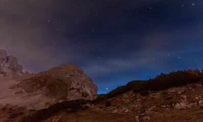 Starry night sky above rugged alpine rocks, calm mountain landscape under deep blue twilight with visible stars and peaceful high altitude atmosphere