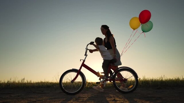 Woman riding bicycle with child on rear seat at sunset in open field, silhouette framed by warm sky, colorful balloon cluster tied to seat, family ride conveying joy and close bond during golden hour
