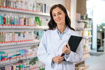 Female pharmacist standing confidently with clipboard near shelves of medicine
