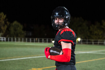 American football player wearing helmet and uniform, holding oval ball on field at night. Displaying sport, strength, determination
