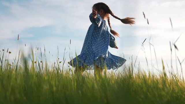 Spinning girl twirling in field of tall grass under clear sky, child wearing blue polka dot dress with braid, arms out, running and play in summer nature, joyful motion with breeze and green grass