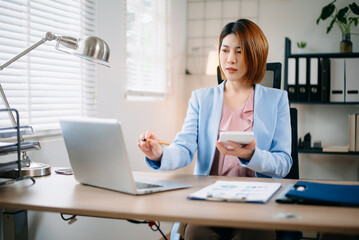 Fototapeta premium Asian businesswoman using calculator and laptop for financial planning in bright office. Ideal for finance, accounting, tax