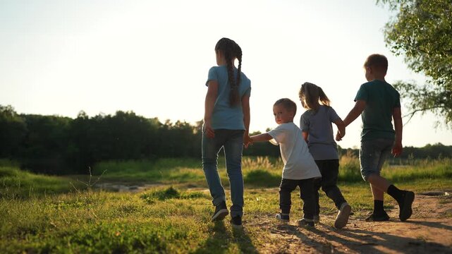 Children walking hand in hand along grassy field at sunset, sunlight lighting child and sibling silhouette while family walk together, friendship visible, nature path, barefoot play, step and movement