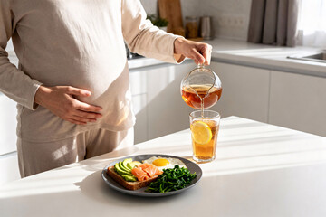 Superfood Breakfast and Lemon Water – Pregnant Woman in Soft Loungewear Pouring Drink in Bright Kitchen, Avocado Toast with Salmon, Greens and Egg on Plate, Copy Space