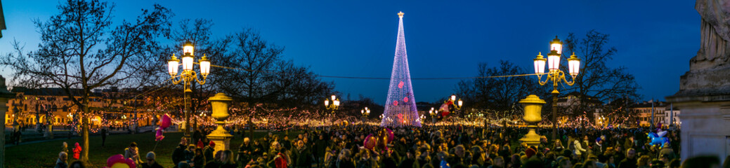 A large crowd gathers in Piazza delle Erbe, Padua, for a Christmas celebration. The square is illuminated by a modern light tree and festive decorations at dusk.