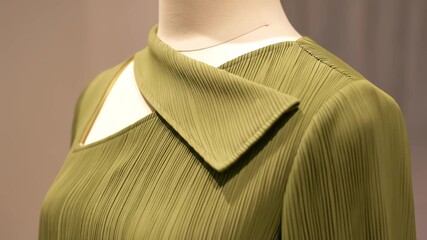 A detailed view of a modern olive green pleated blouse on a mannequin, featuring a unique asymmetrical neckline with a zipper