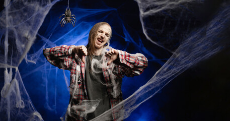Dead guy with horrible scary Halloween zombie makeup on face showing thumb down dislike gesture standing covered in spiderwebs on blue background