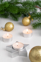 Christmas decorations with glowing  candles light in star shape silver holder, green fir tree branches and golden glittering ornament baubles on white wooden table prepared for cozy winter holidays