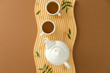White ceramic teapot and cups on wavy wood tray with green leaves, styled on brown surface
