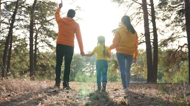 Family walking hand in hand along forest trail under golden sunlight with parent mother and father supporting child between pine tree trunks wearing casual clothes showing family joy and nature bond