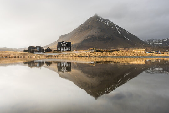 View of a dark, imposing house stands starkly against the golden, windswept landscape, mirrored perfectly in the still waters beneath a majestic mountain, Iceland.