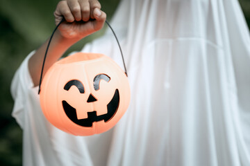 A young boy dressed as a ghost for Halloween wears a white sheet with black eye holes. Holding a pumpkin jack-o’-lantern bucket, he stands outdoors surrounded by dark green foliage