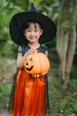 A cute little girl smiles brightly, dressed as a witch for Halloween. She wears a black pointed hat, an orange-and-black costume, holding a jack-o&rsquo;-lantern pumpkin bucket outdoors among green trees