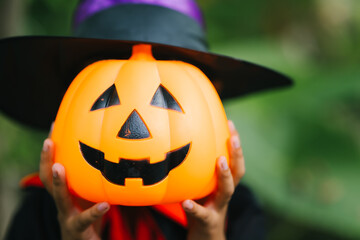 A cheerful young girl dressed in a Halloween witch costume smiles happily outdoors. Wearing a pointed black hat and orange dress, she holds a pumpkin jack-o’-lantern bucket, surrounded by greenery