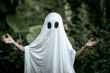 A young boy dressed as a ghost for Halloween wears a white sheet with two black eye holes. He raises both arms in a spooky gesture, surrounded by dark green foliage
