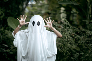 A young boy dressed as a ghost for Halloween wears a white sheet with two black eye holes. He raises both arms in a spooky gesture, surrounded by dark green foliage