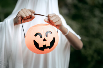 A young boy dressed as a ghost for Halloween wears a white sheet with black eye holes. Holding a pumpkin jack-o’-lantern bucket, he stands outdoors surrounded by dark green foliage