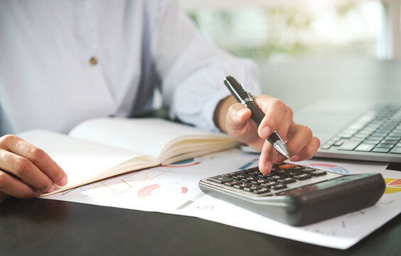 Businessperson using calculator with financial charts and laptop on desk office