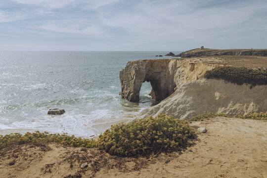 View of rugged cliffs  of Arche De Port Blanc Roche meet the turquoise sea, carving out natural arches beneath a pale sky, a lighthouse stands guard, bathed in the golden hues, Brittany, France.